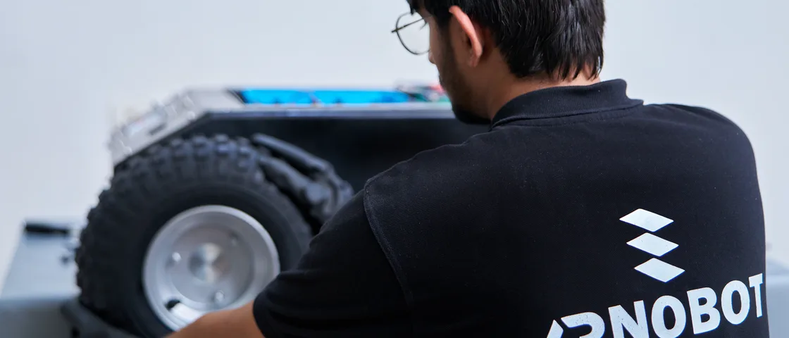 An ARNOBOT engineer in a branded polo refitting a drive wheel to a UGV chassis on the bench, the battery pack open on the deck behind.